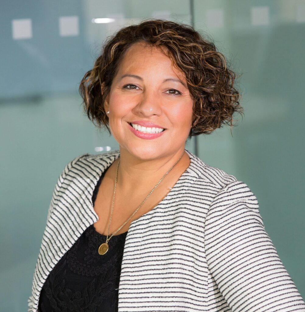 A confident woman with curly hair smiling in an office environment, conveying professionalism and approachability.